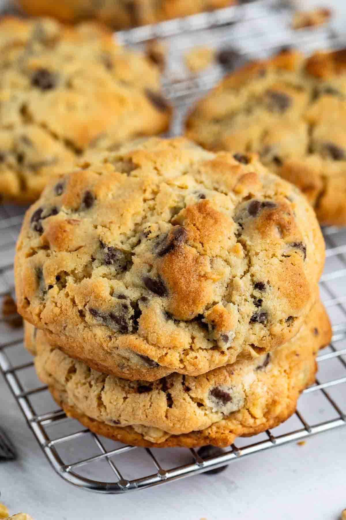 Two large, golden-brown chocolate chip cookies are stacked on a wire cooling rack, with more cookies blurred in the background. The cookies have a chunky, crumbly texture and visible chocolate chips.