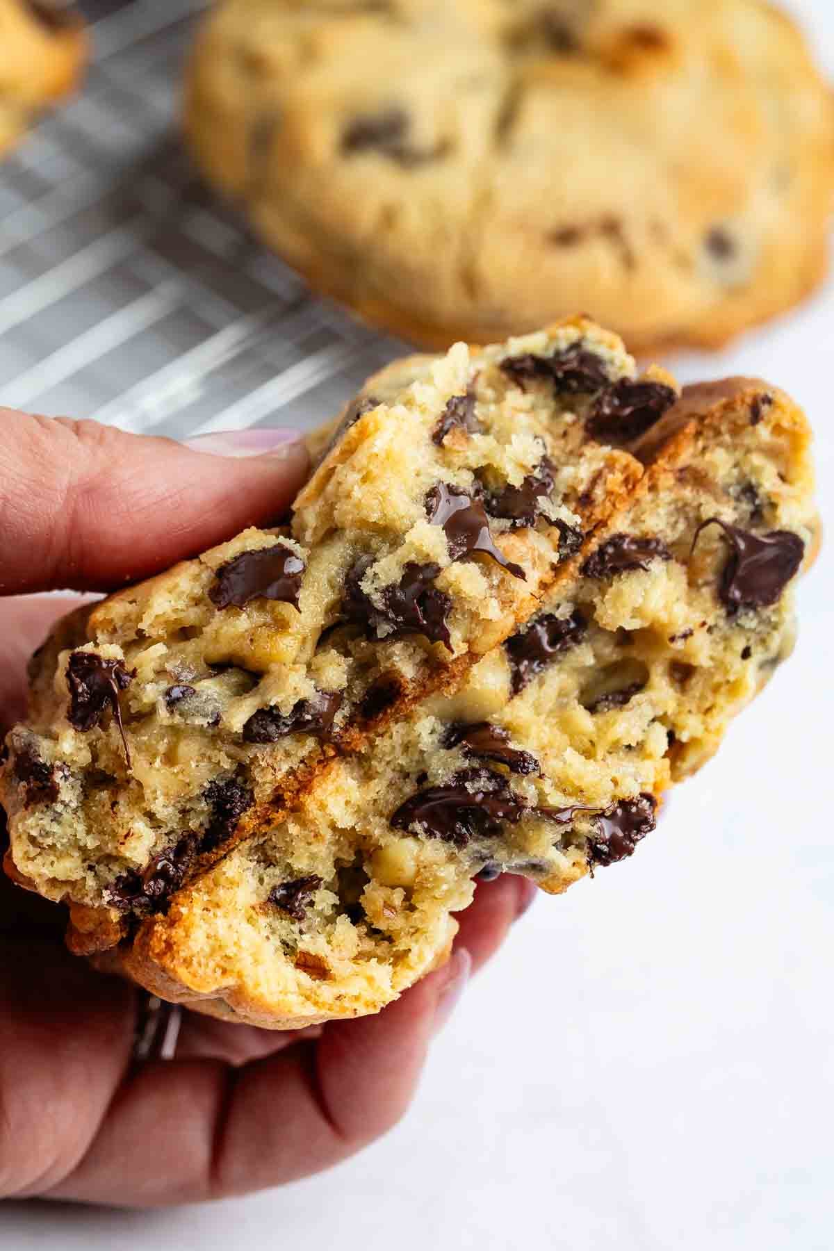 A hand holds a thick, gooey chocolate chip cookie broken in half, revealing a soft, melty interior with chunks of chocolate. A wire cooling rack and more cookies are blurred in the background.