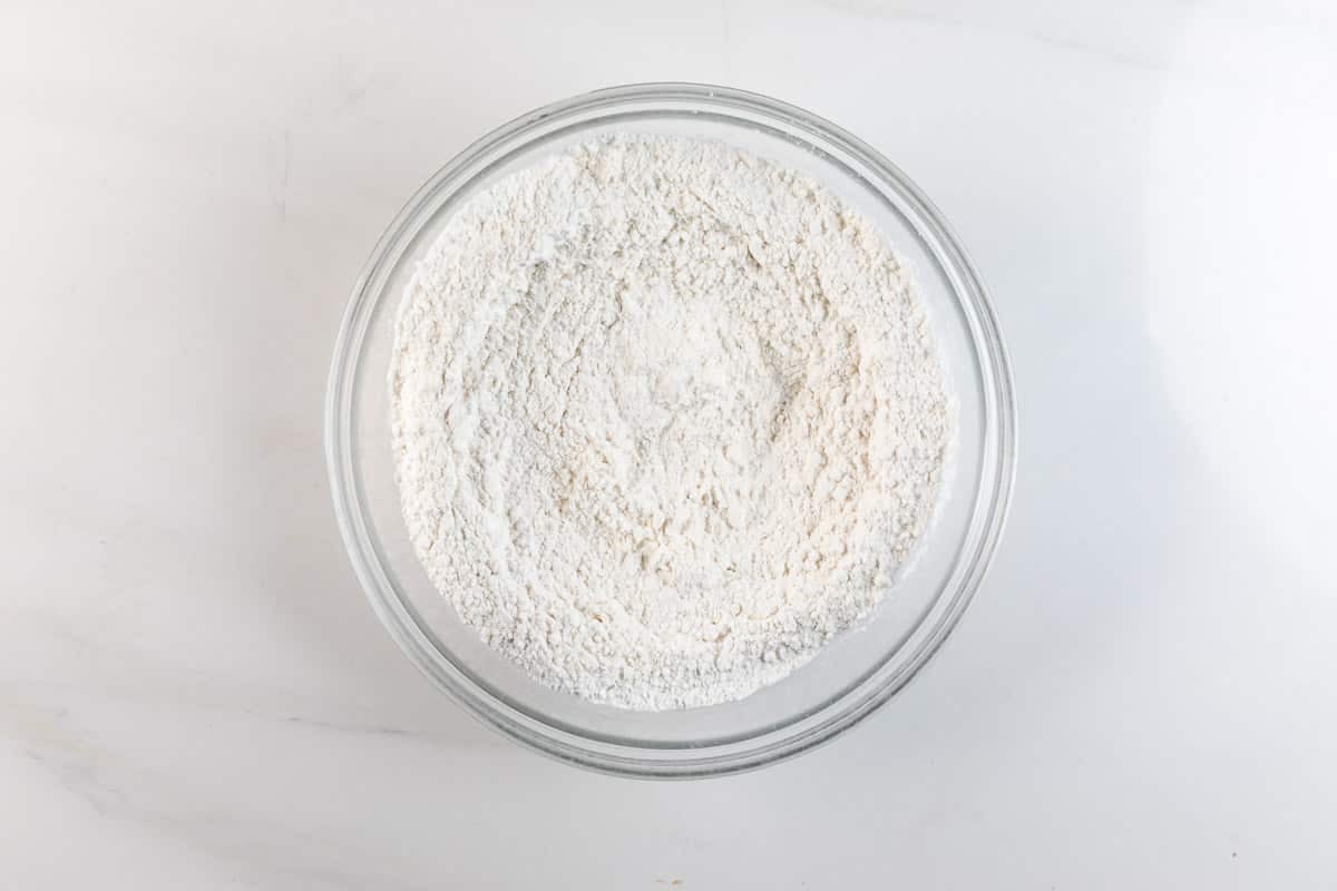 A clear glass bowl filled with white flour sits on a white marble surface.
