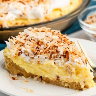 A slice of coconut cream pie topped with toasted shredded coconut sits on a white plate with a fork, with the rest of the pie and a small bowl of coconut in the background.