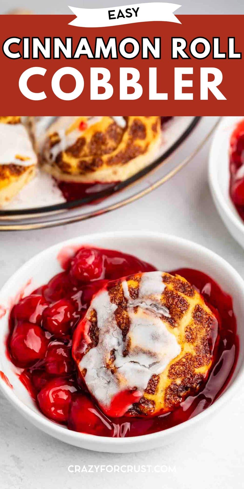 A white bowl filled with cherry cobbler topped with a cinnamon roll and white icing. In the background, there is a plate with more cobbler. Text reads Easy Cinnamon Roll Cobbler at the top.