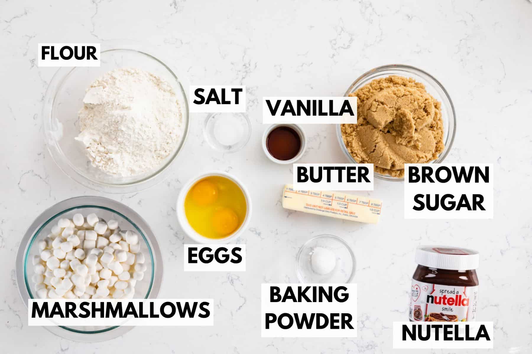 Top-down view of baking ingredients on a white surface, labeled: flour, salt, vanilla, butter, brown sugar, Nutella, baking powder, eggs, and marshmallows, each in bowls or their original containers.