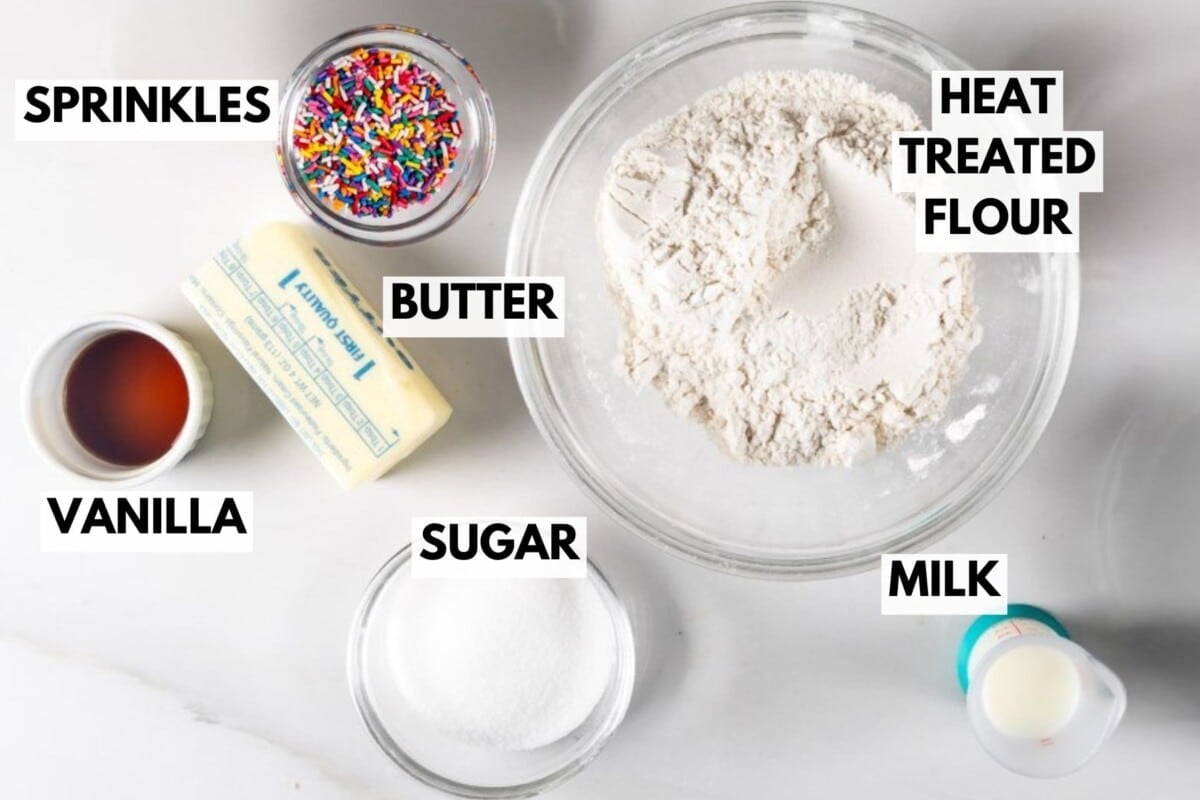 A top-down view of ingredients in bowls and containers labeled as sprinkles, butter, sugar, vanilla, milk, and a large bowl of heat-treated flour on a white surface.