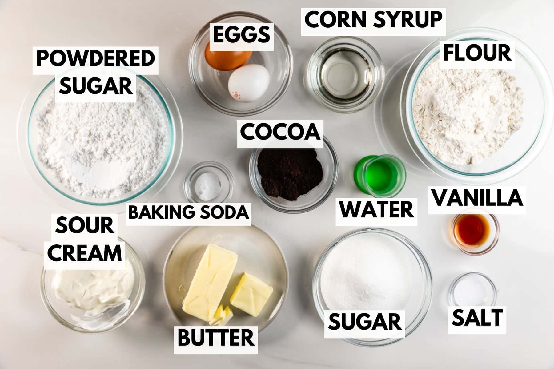 Une vue de haut en bas des ingrédients de cuisson présentés dans des bols et des assiettes, notamment du sucre en poudre, des œufs, du sirop de maïs, de la farine, du cacao, de la vanille, de l'eau, du sucre, du sel, de la crème sure, du beurre et du bicarbonate de soude, disposés sur une surface blanche.