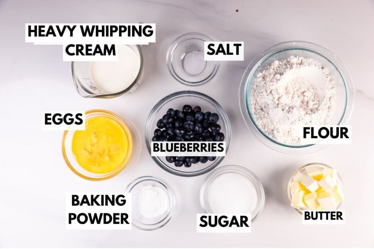 Top-down view of labeled baking ingredients for moist blueberry scones in bowls: heavy whipping cream, eggs, blueberries, baking powder, salt, flour, sugar, and cubed butter—all neatly arranged on a white surface.