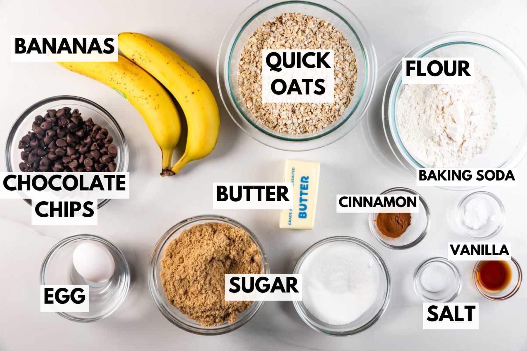Vue de dessus des ingrédients de cuisson affichés sur une surface blanche : banane, avoine instantanée, farine, pépites de chocolat, beurre, cannelle, vanille, bicarbonate de soude, sel, sucre et œuf.
