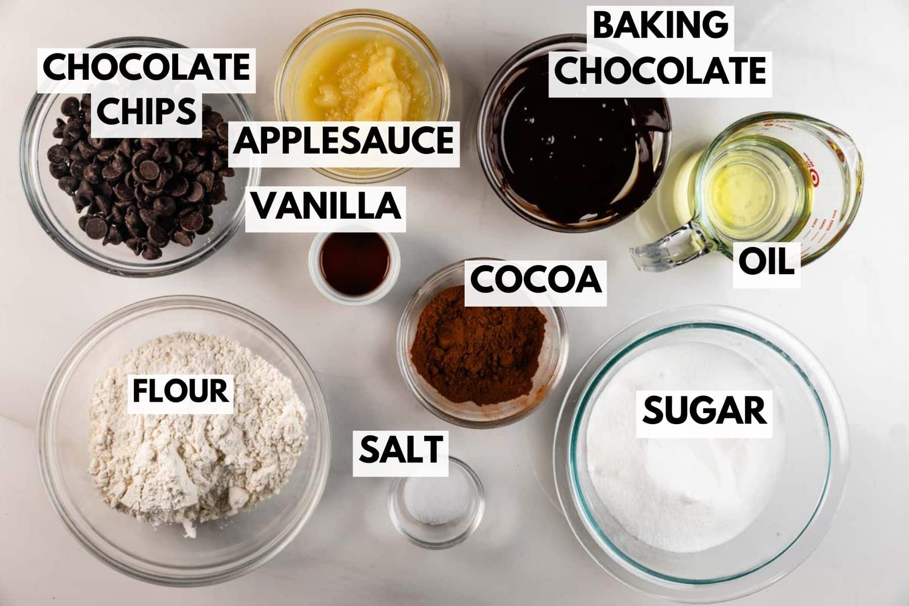 Top-down view of baking ingredients in bowls labeled: chocolate chips, applesauce, vanilla, flour, salt, cocoa, sugar, oil, and baking chocolate, all arranged on a white surface.