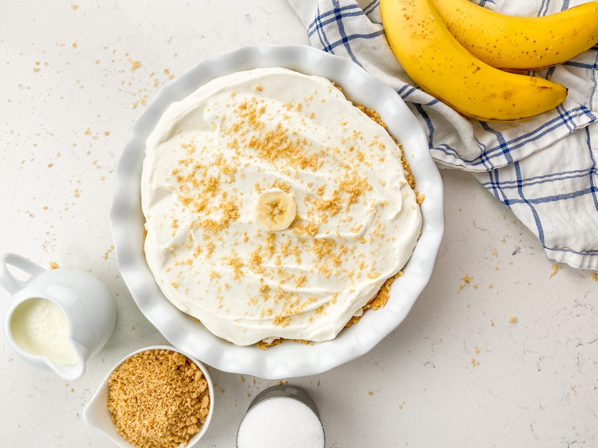 A creamy pie topped with a banana slice and crumbled crumbs sits on a white counter. Nearby are bananas, a blue-and-white towel, a cup of milk, a bowl of crumbs, and a sugar jar.