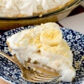 A slice of banana cream pie with whipped topping and banana slices on a blue and white patterned plate, with a fork beside it and the rest of the pie in the background.