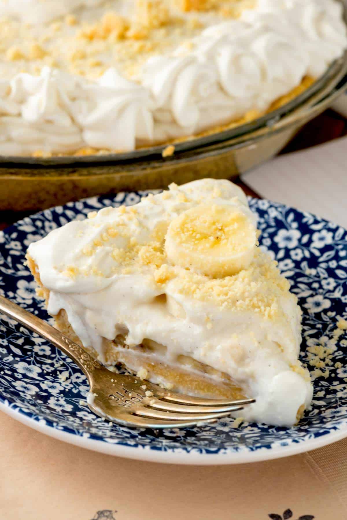 A slice of banana cream pie with whipped topping and banana slices on a blue and white patterned plate, with a fork beside it and the rest of the pie in the background.