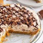 A creamy, chocolate-drizzled pie with a graham cracker crust, topped with chopped chocolate pieces, sits in a glass pie dish with a large slice missing.