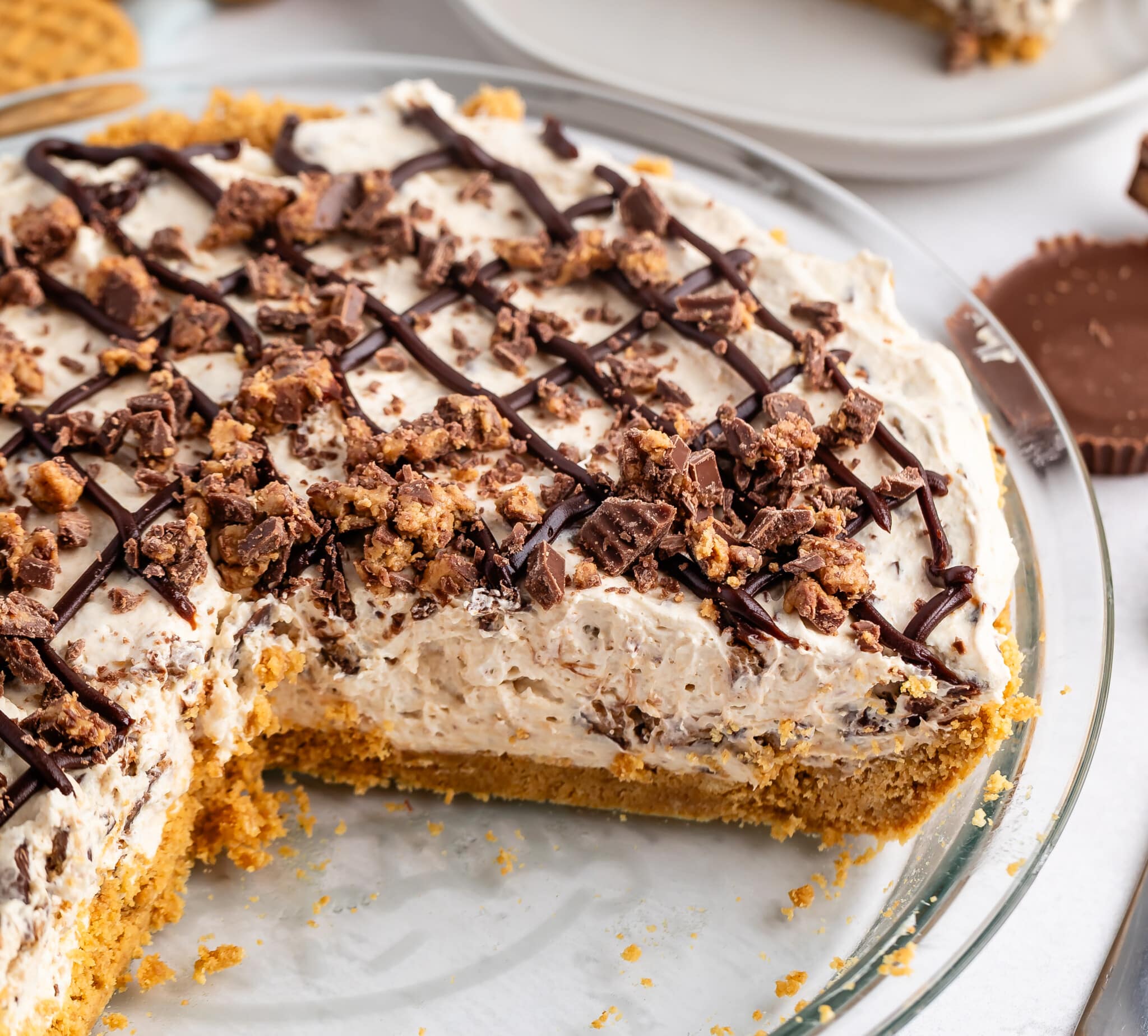 A creamy, chocolate-drizzled pie with a graham cracker crust, topped with chopped chocolate pieces, sits in a glass pie dish with a large slice missing.
