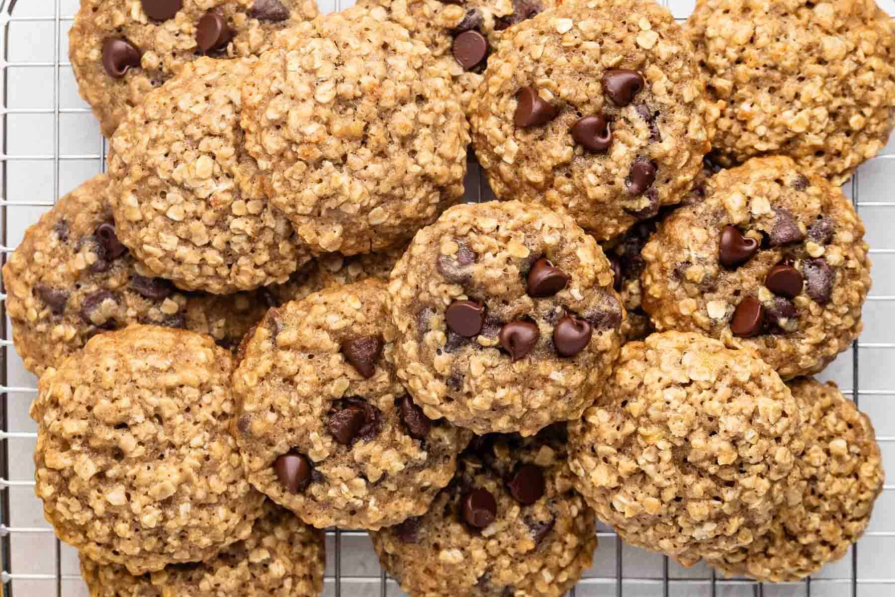 Une pile de biscuits aux pépites de chocolat et à l'avoine empilés sur une grille de refroidissement en fil métallique, avec de l'avoine et des pépites de chocolat visibles partout sur les biscuits dorés.