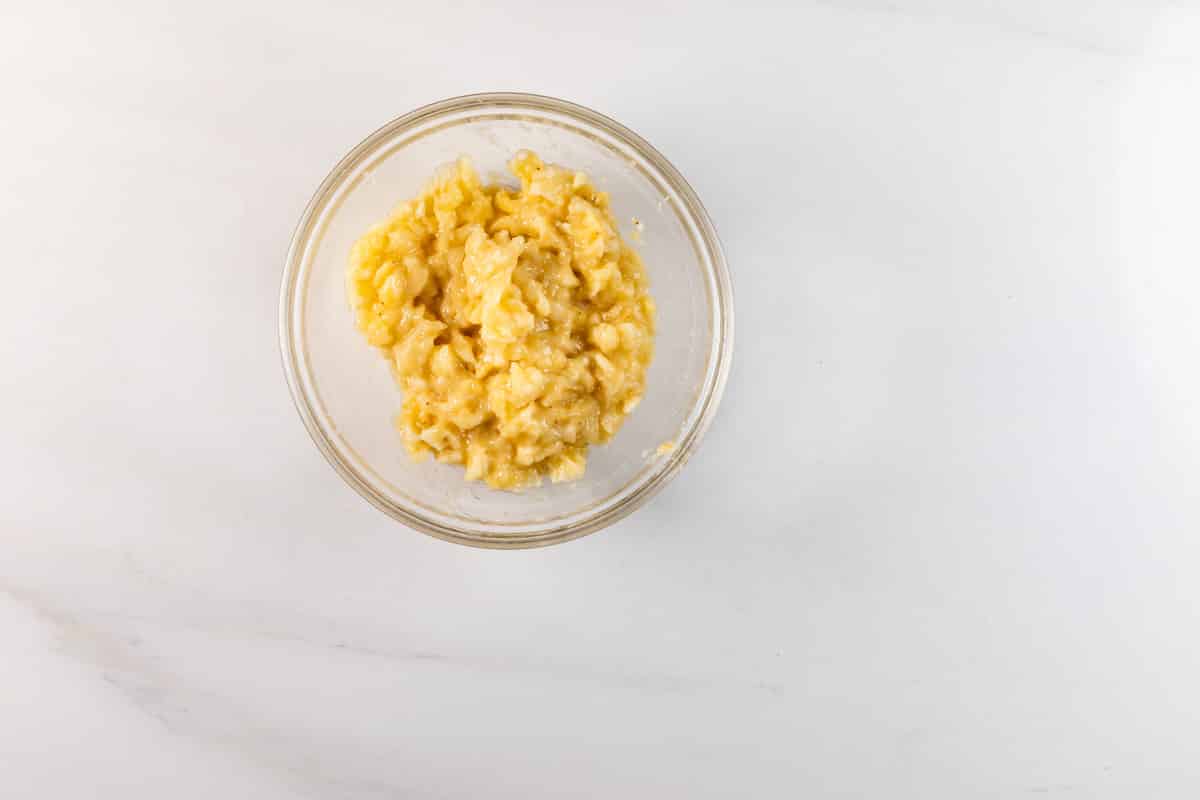 Un bol en verre transparent rempli de purée de bananes repose sur une surface en marbre blanc, légèrement à gauche du centre de l'image.