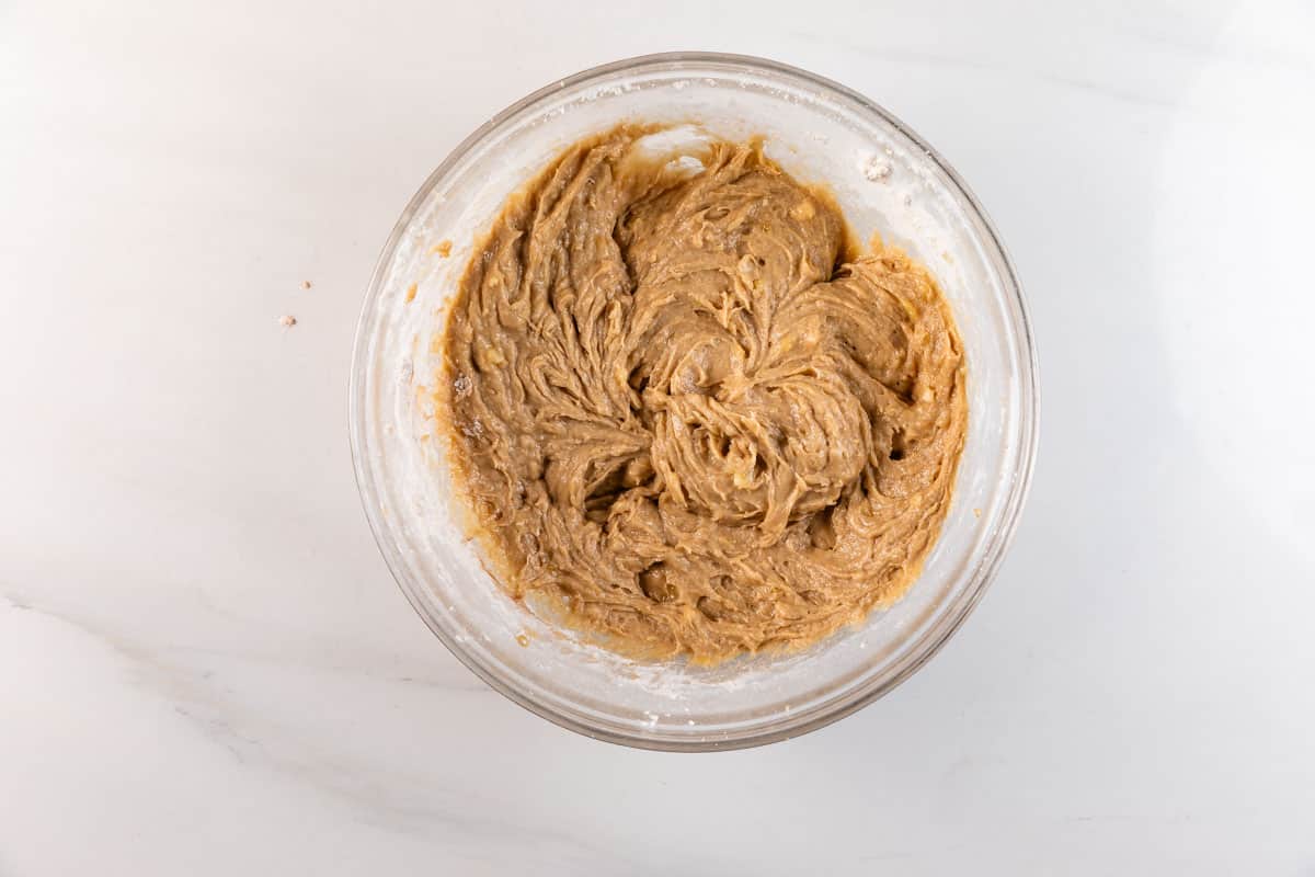 Un bol en verre rempli de mélange de pâte à biscuits brun clair sur une surface en marbre blanc, vue d'en haut.