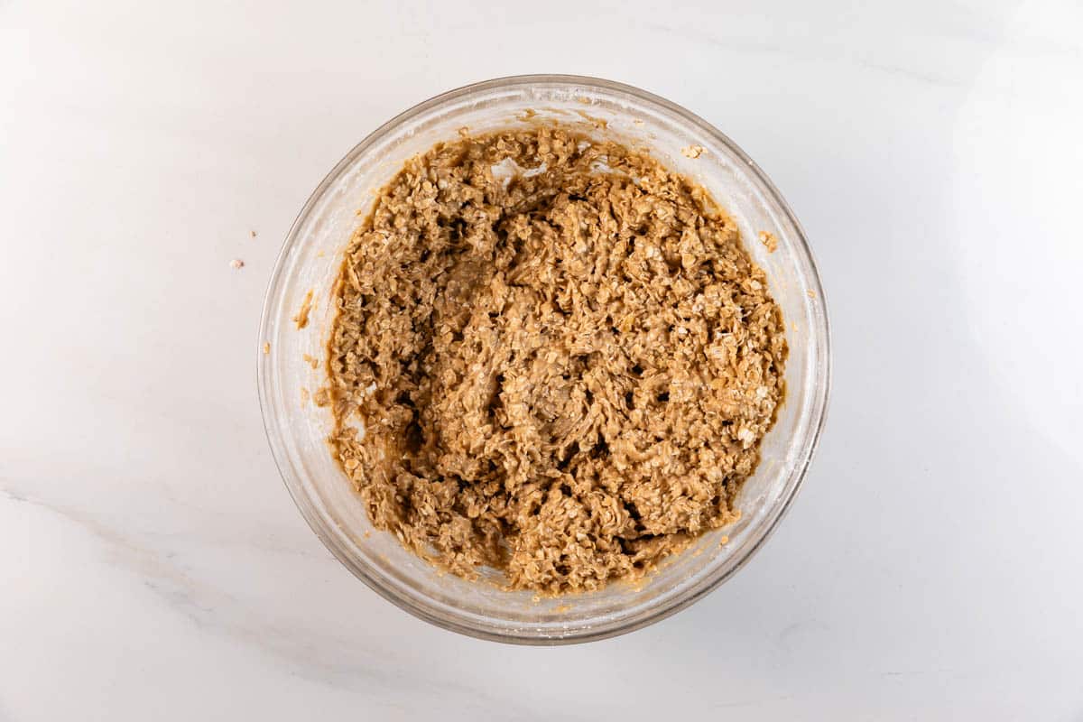 Un bol en verre rempli d'un épais mélange de pâte à biscuits à l'avoine, placé sur un comptoir en marbre blanc.