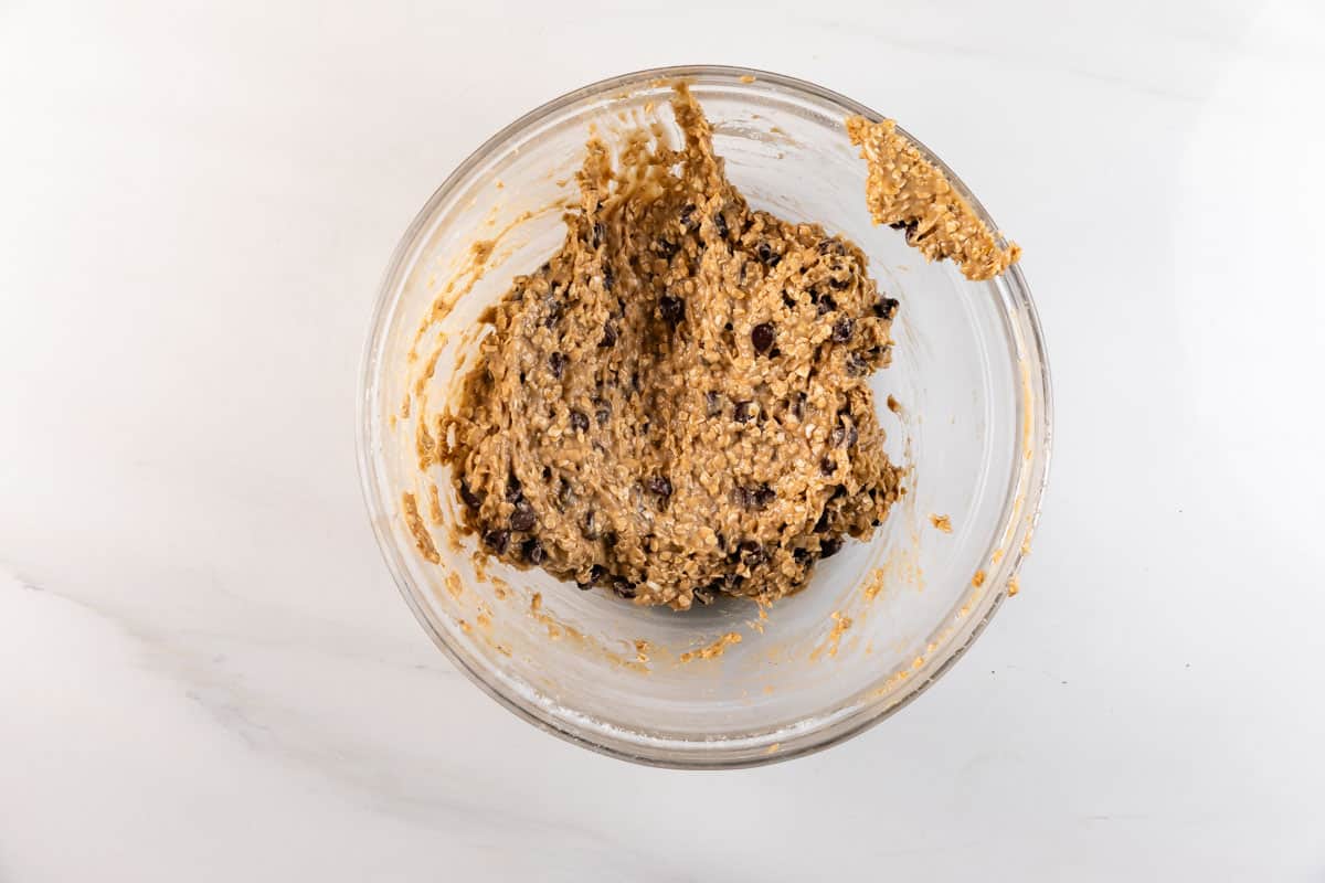 Un bol en verre contenant une épaisse pâte à biscuits à l'avoine avec des pépites de chocolat visibles, placé sur une surface de travail blanche.