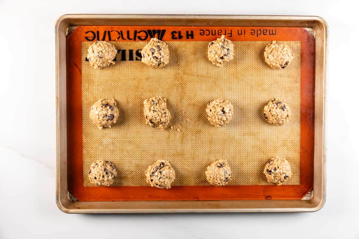 Une plaque à pâtisserie tapissée de silicone contient douze boules de pâte à biscuits crues uniformément espacées, prêtes à cuire. Les biscuits contiennent des pépites de chocolat et d'avoine visibles.