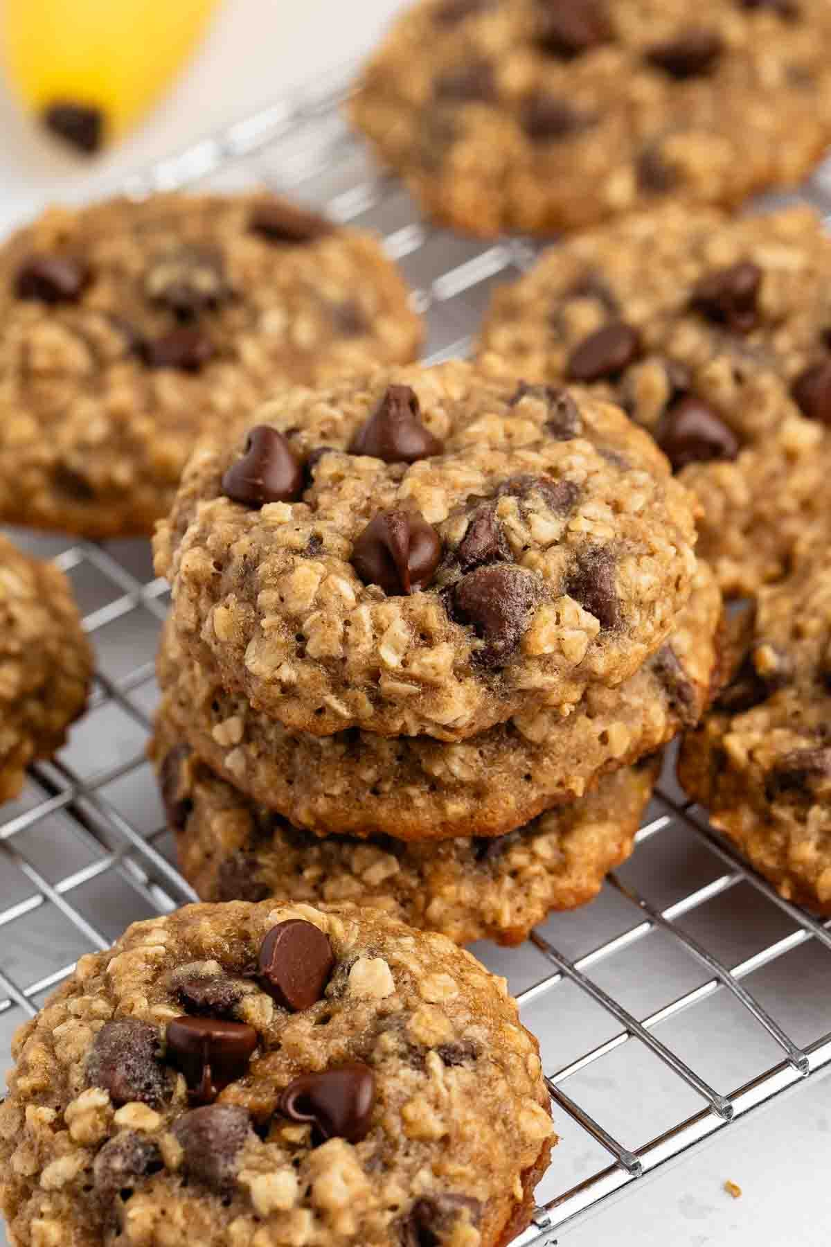 Une pile de biscuits aux pépites de chocolat et à l'avoine faits maison se trouve sur la grille de refroidissement, avec d'autres biscuits et une banane floue en arrière-plan. Les biscuits sont dorés et parsemés de pépites de chocolat.
