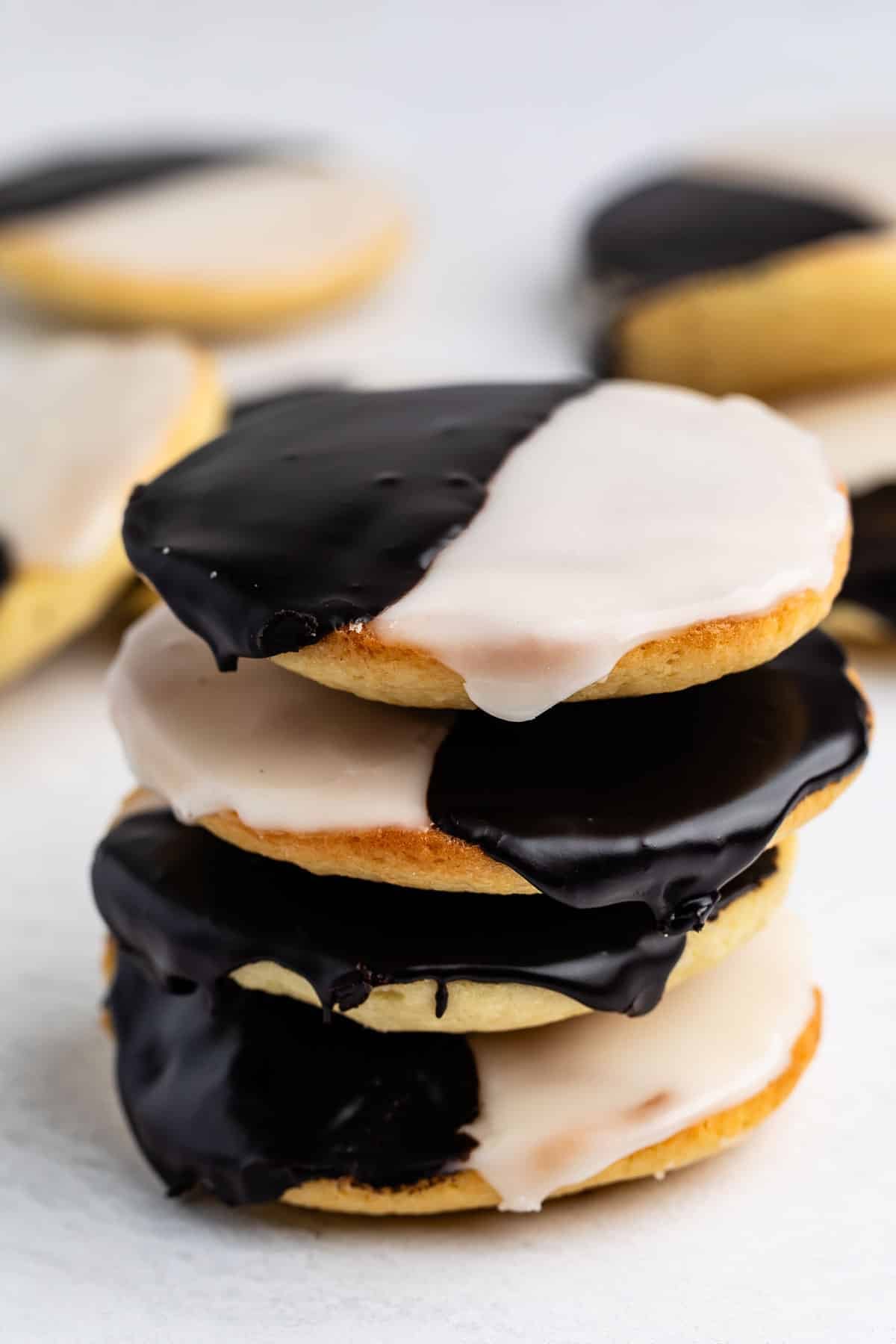 Une pile de trois biscuits noirs et blancs, chaque moitié recouverte d'un glaçage au chocolat et l'autre moitié d'un glaçage blanc, placée sur une surface blanche avec d'autres biscuits flous en arrière-plan.