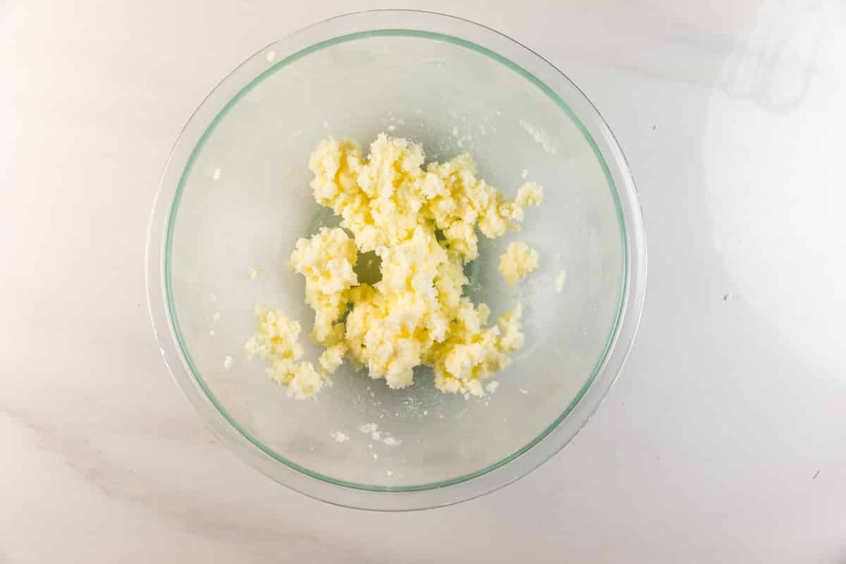 Un bol en verre contenant un mélange de beurre et de sucre posé sur une surface blanche.