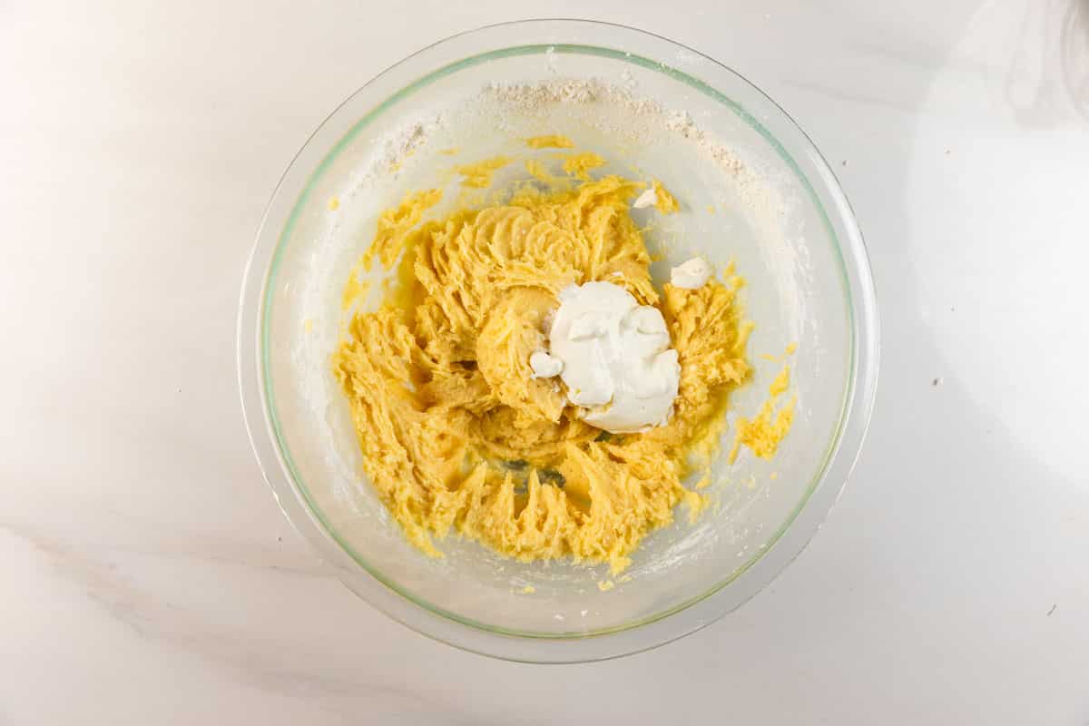 Un bol à mélanger en verre avec une pâte à gâteau jaune et une cuillerée de crème sure blanche sur le dessus, placé sur une surface de travail blanche.