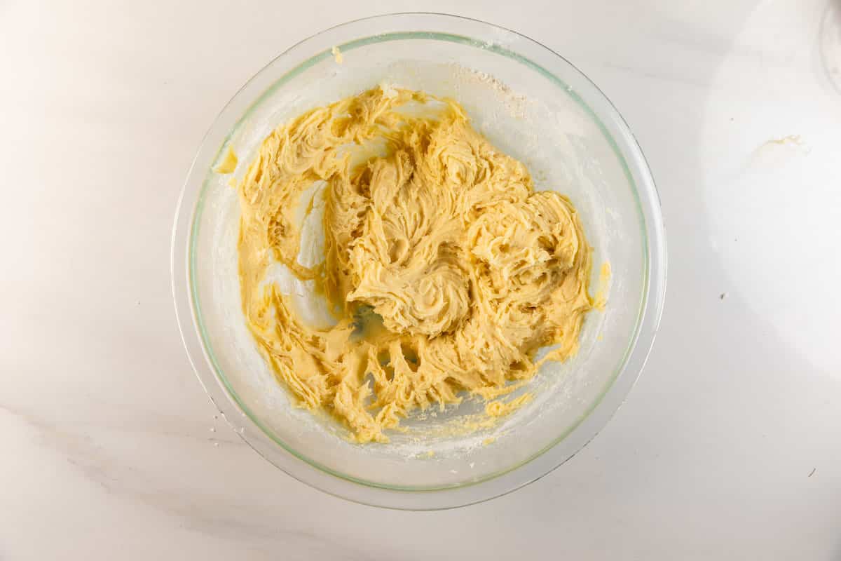 Un bol à mélanger en verre contenant une pâte à biscuits crémeuse jaune clair repose sur un plan de travail blanc. La pâte semble bien mélangée et légèrement collante.