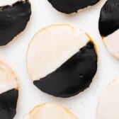 Round cookies topped with half white icing and half black icing are arranged on a white surface. The cookies are known as black and white cookies.