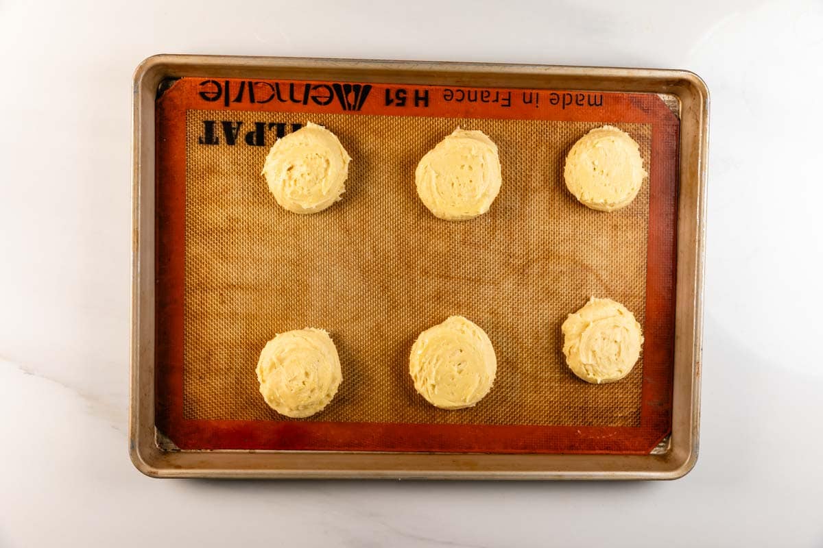 Une plaque à pâtisserie recouverte d'un tapis en silicone contient six boules de pâte à biscuits uniformément espacées, prêtes à cuire. Posé sur un comptoir blanc.