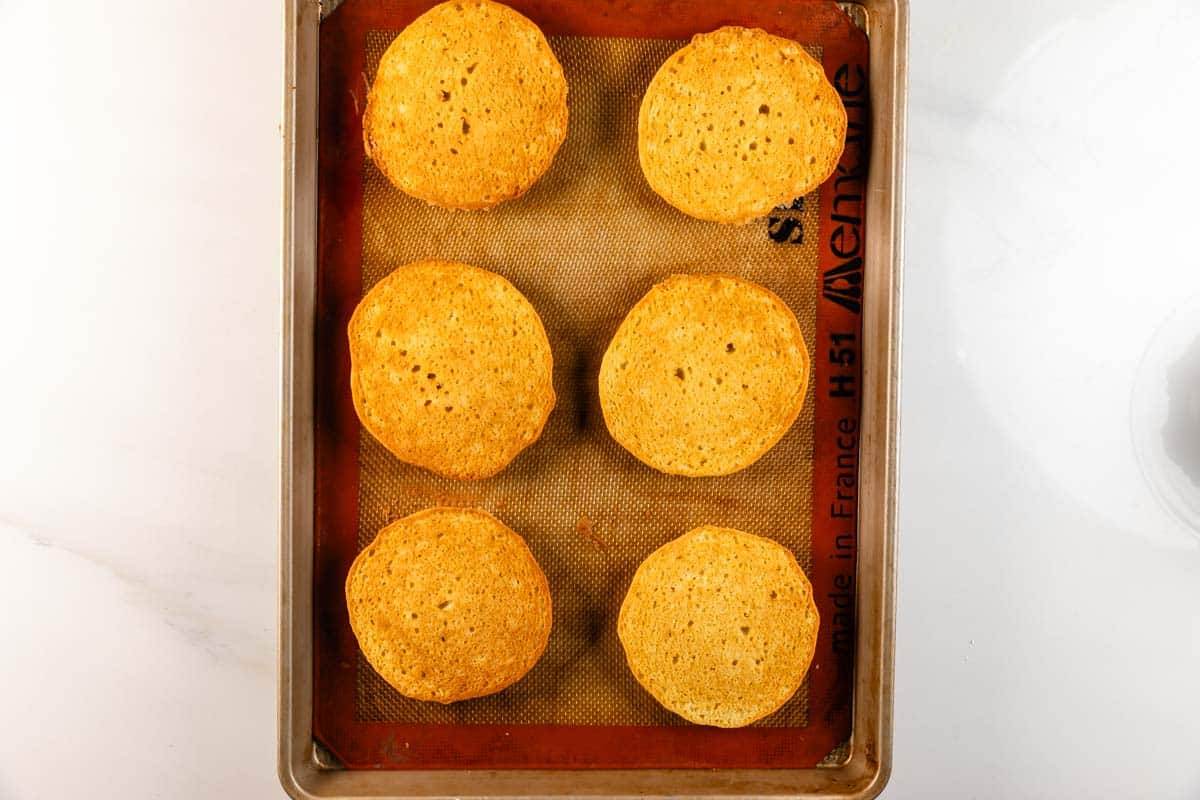 Une plaque à pâtisserie recouverte d'un tapis de cuisson en silicone contient six biscuits brun doré uniformément arrondis sur une surface de travail blanche.