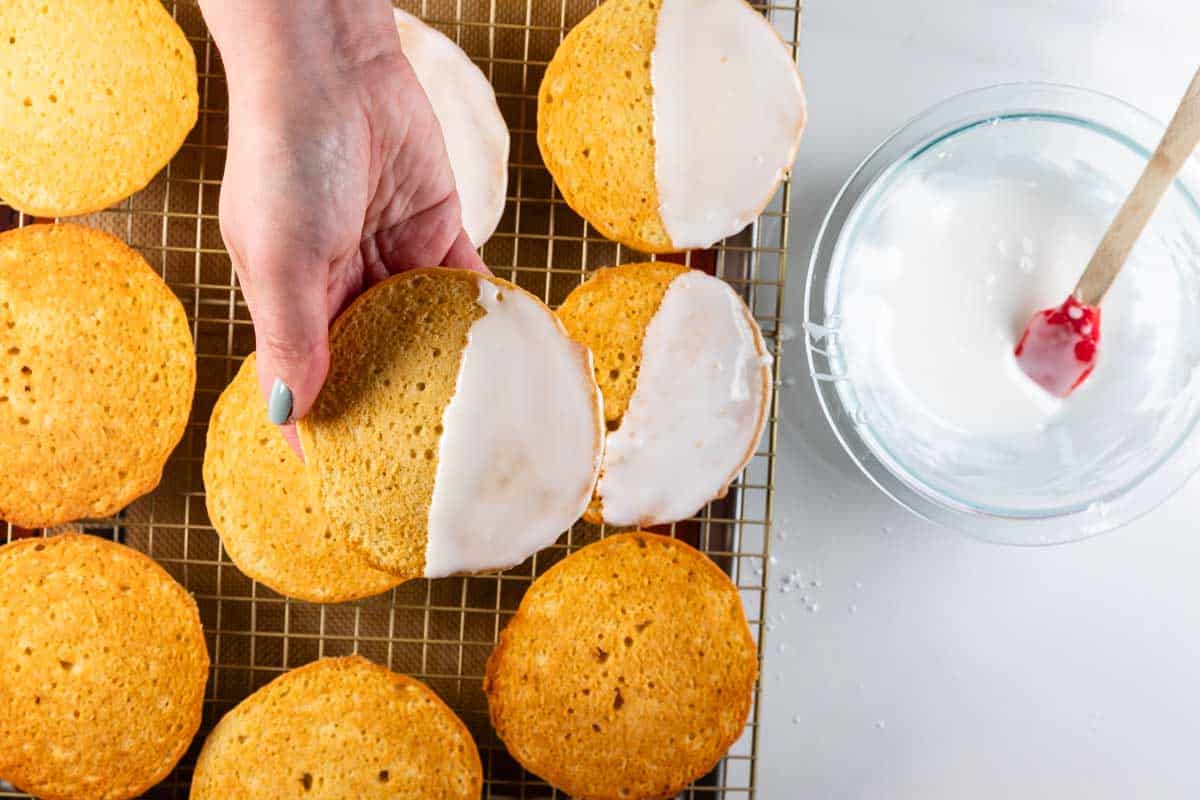 La main tient un gâteau rond à moitié trempé avec un glaçage blanc sur une grille de refroidissement avec des biscuits assortis. Il y a un bol en verre de crème blanche avec une cuillère rouge à proximité.