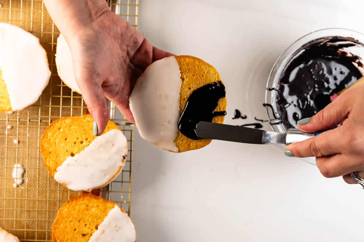 Une personne étale une couche de chocolat sur la moitié d’un biscuit tout en recouvrant l’autre moitié d’une couche de glaçage blanc. Placez les biscuits réfrigérés sur une grille de refroidissement à proximité.