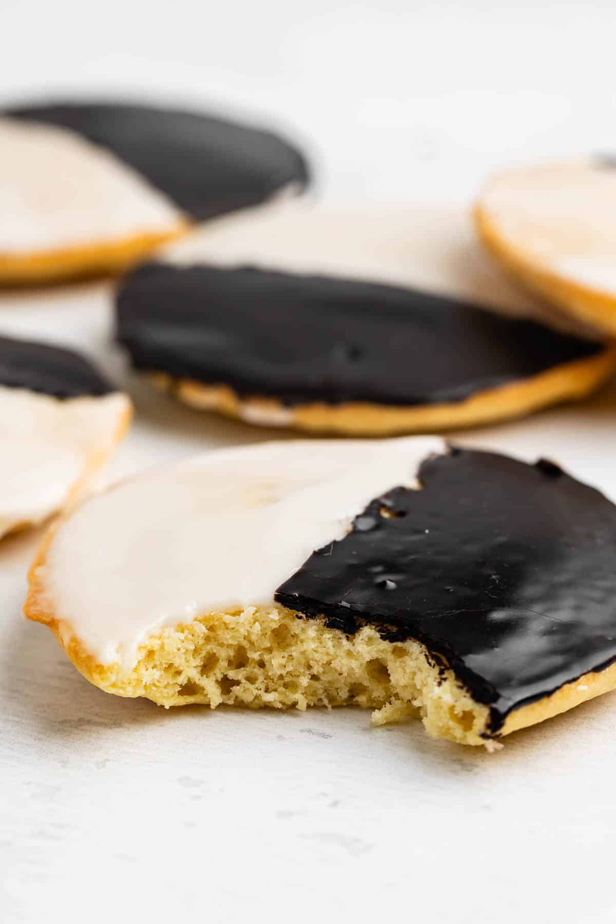 Gros plan sur un biscuit noir et blanc, dont un au premier plan sans bouchée. La moitié des biscuits est recouverte d'un glaçage blanc et l'autre moitié d'un glaçage au chocolat, et déposée sur une surface blanche.