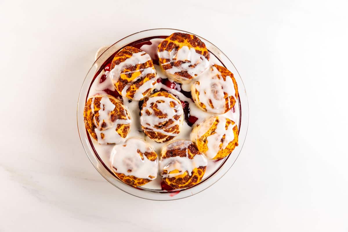A round baking dish filled with golden-brown cinnamon rolls topped with white icing, arranged on a white surface. Some of the rolls show hints of red filling peeking through.
