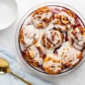 A round baking dish filled with cinnamon rolls topped with icing, next to a white bowl, a gold spoon, and a light blue napkin on a white surface.