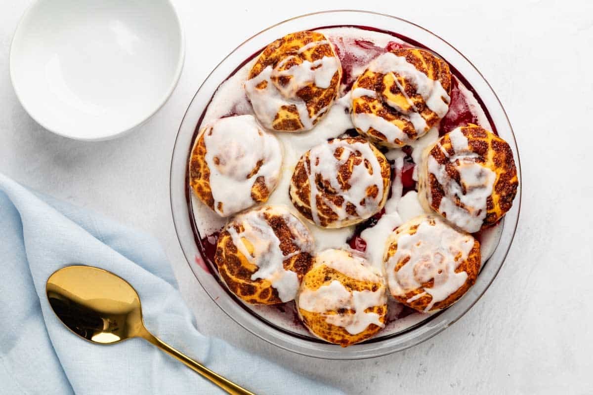 A round baking dish filled with cinnamon rolls topped with icing, next to a white bowl, a gold spoon, and a light blue napkin on a white surface.