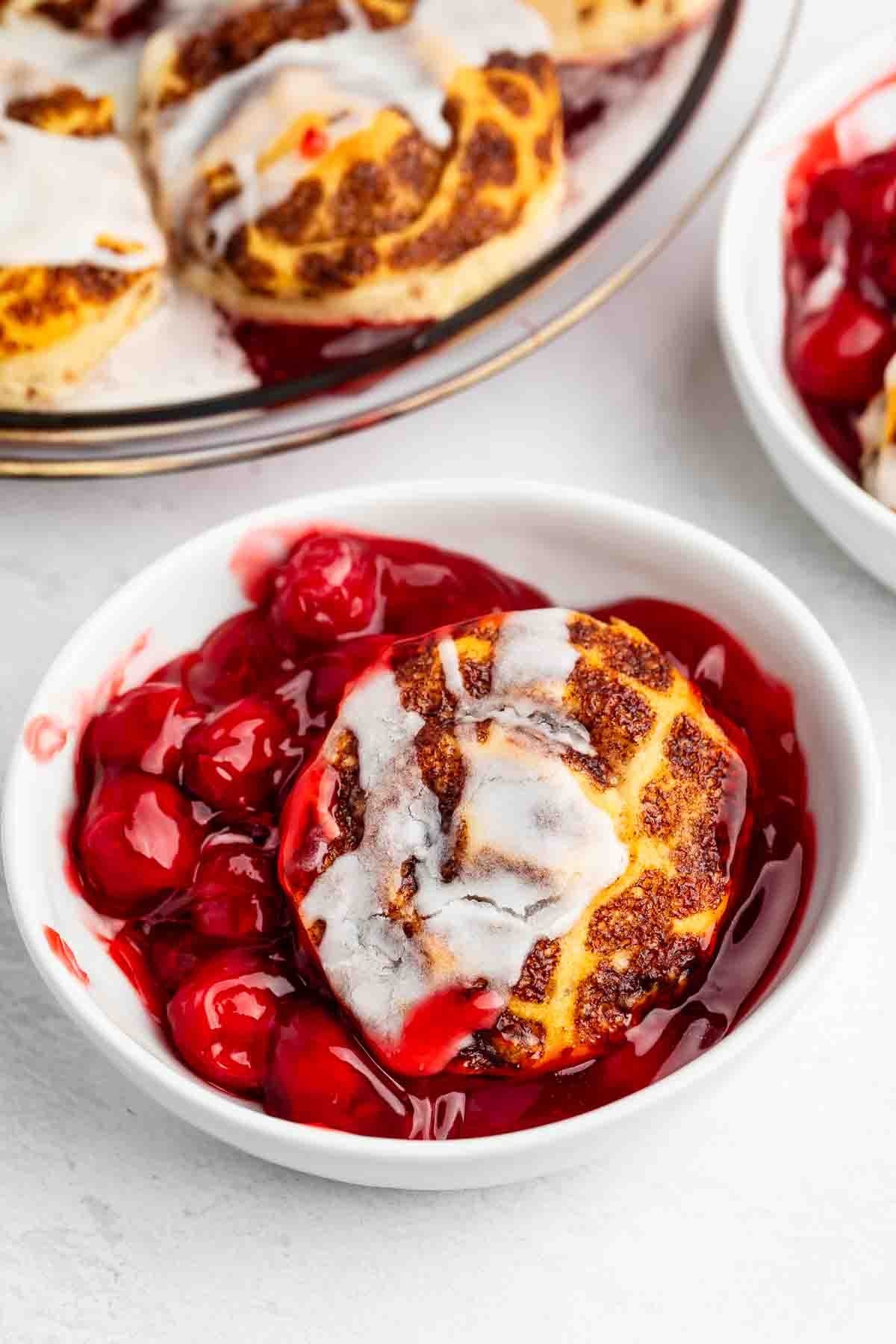 A white bowl filled with cherry pie filling and a golden-brown biscuit topped with white icing. Another plate with more biscuits and cherries is partially visible in the background.