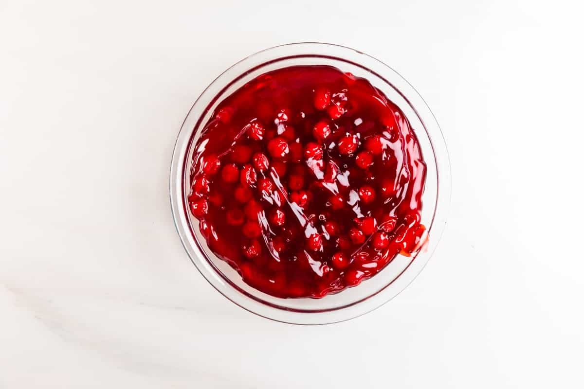 A clear glass bowl filled with bright red cherry pie filling, showing whole cherries and glossy syrup, sits on a white marble surface.