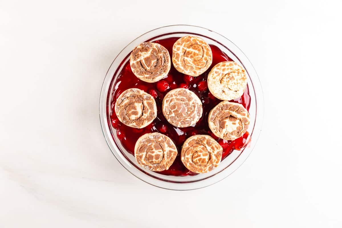A glass pie dish filled with bright red cherry filling, topped with nine round, golden-brown biscuit pieces arranged in a circular pattern. The dish is on a white surface.
