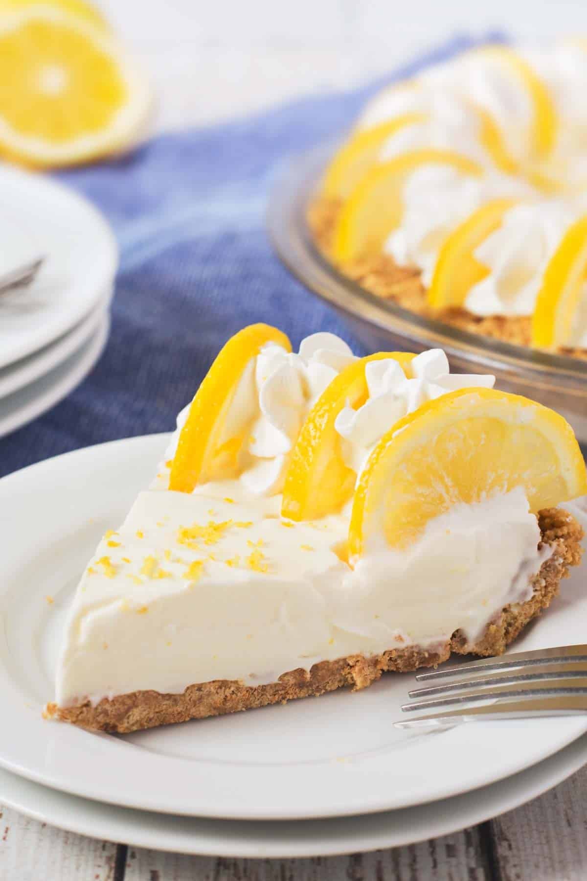 A slice of creamy lemon pie with a graham cracker crust, topped with whipped cream and fresh lemon slices, sits on a white plate with a fork. The rest of the pie is visible in the background.