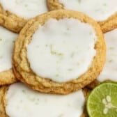 A close-up of several cookies with a light glaze on top, showing flecks of lime zest, arranged on a plate with a sliced lime nearby.