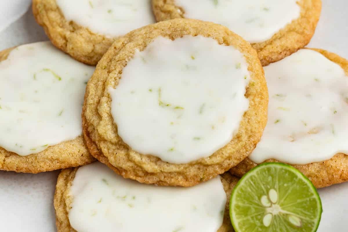 A close-up of several cookies with a light glaze on top, showing flecks of lime zest, arranged on a plate with a sliced lime nearby.