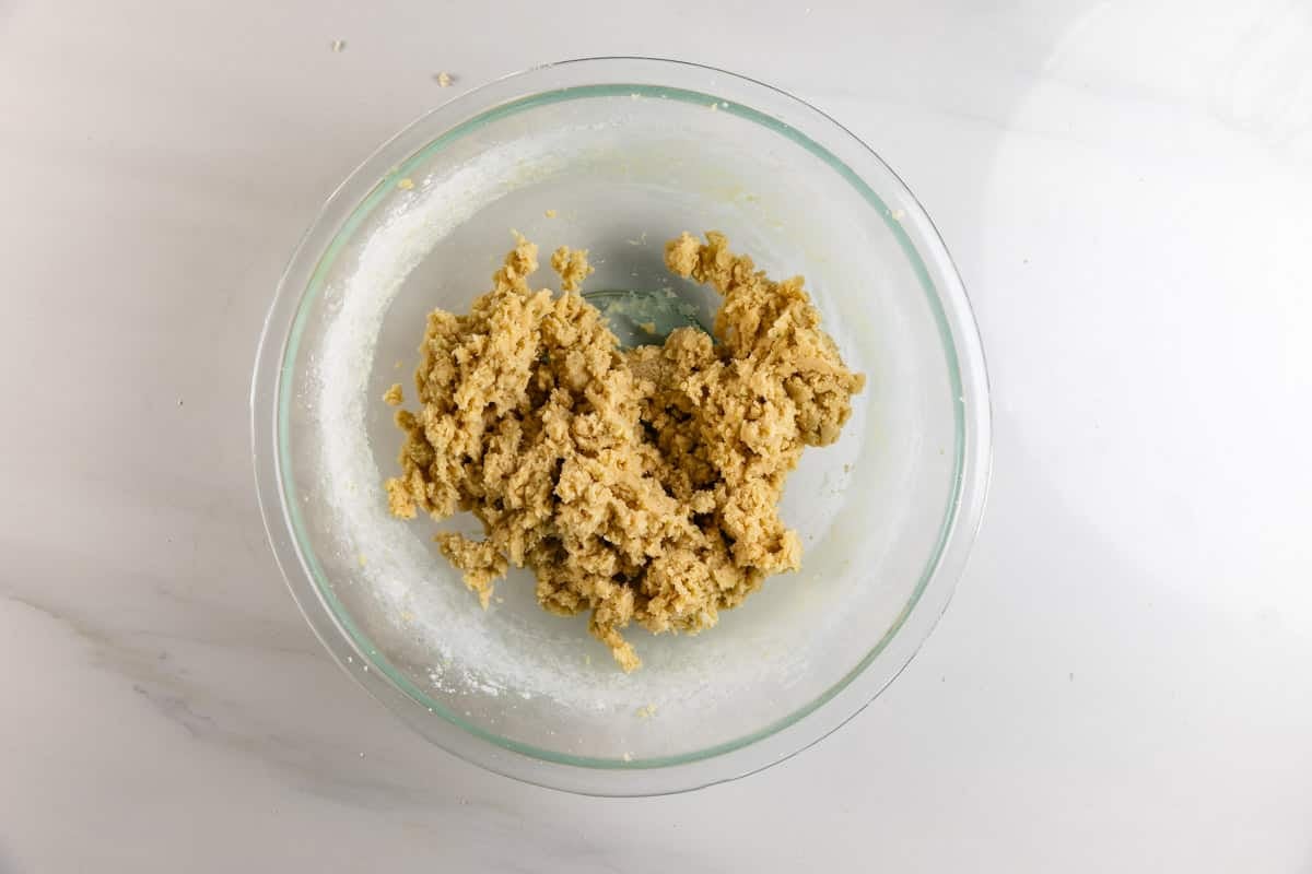 A glass bowl containing a rough, beige cookie dough mixture sits on a white marble surface.