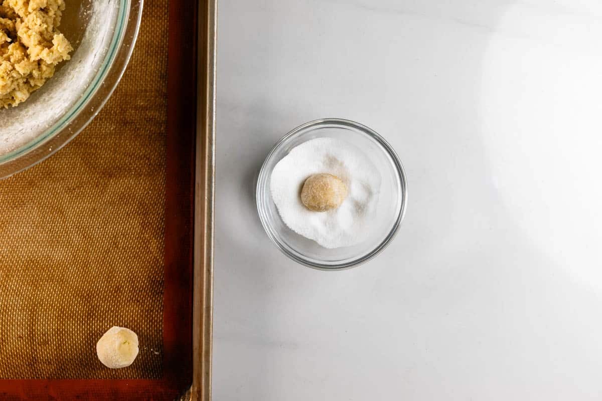A small ball of cookie dough sits in a bowl of white sugar, next to a baking sheet with a silicone mat and another dough ball, with a mixing bowl partially visible at the top left.
