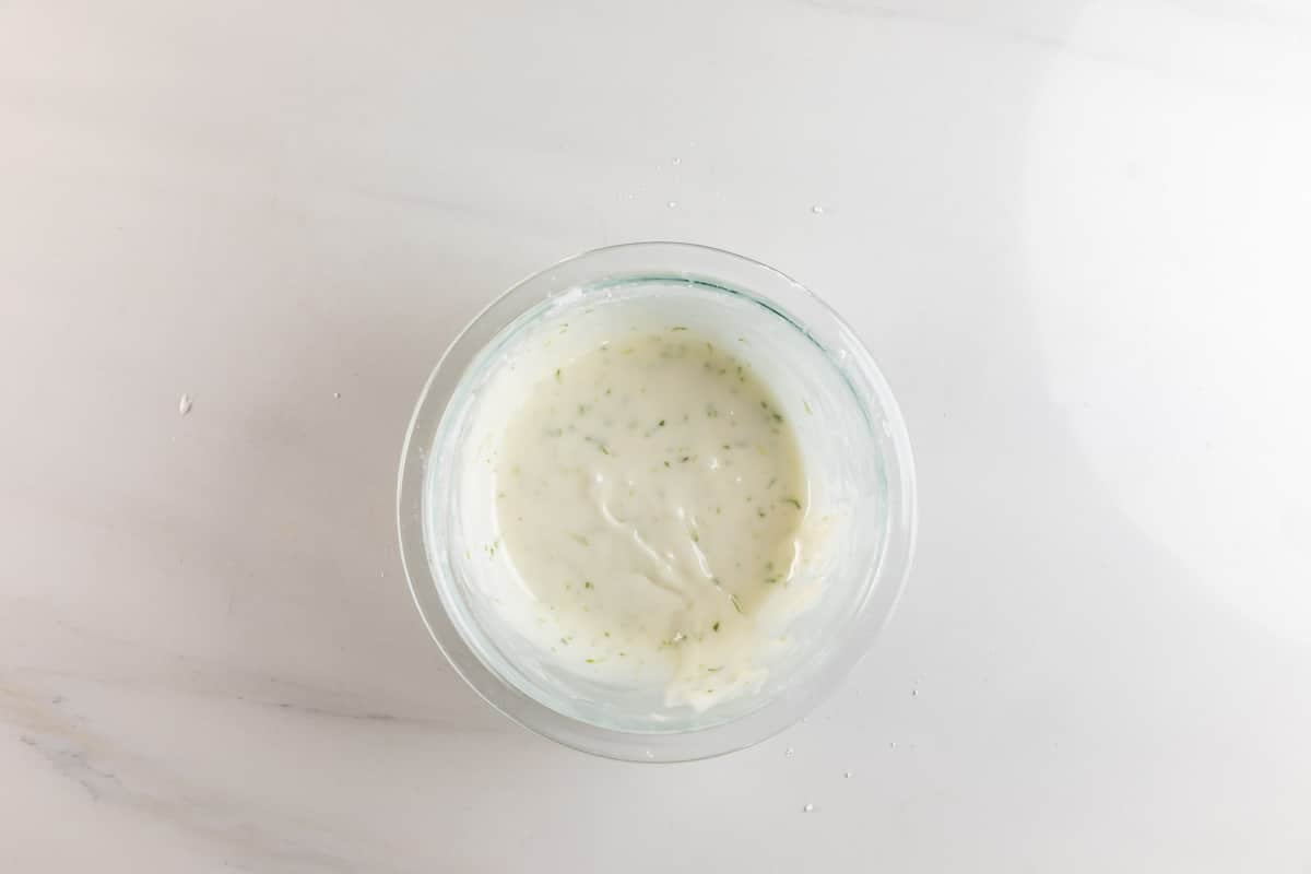 A glass bowl filled with creamy white sauce, speckled with green herbs, sits on a white marble surface.
