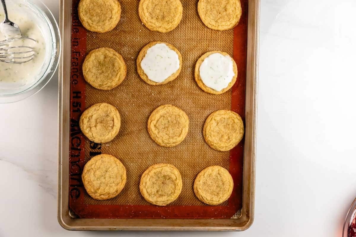 A baking sheet with round cookies on a silicone mat; two cookies have white frosting spread on top, and a glass bowl with a metal whisk and more frosting is in the upper left corner.