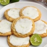 A plate of five cookies with white icing and lime zest, arranged around a sliced lime. More cookies and whole limes are in the background.