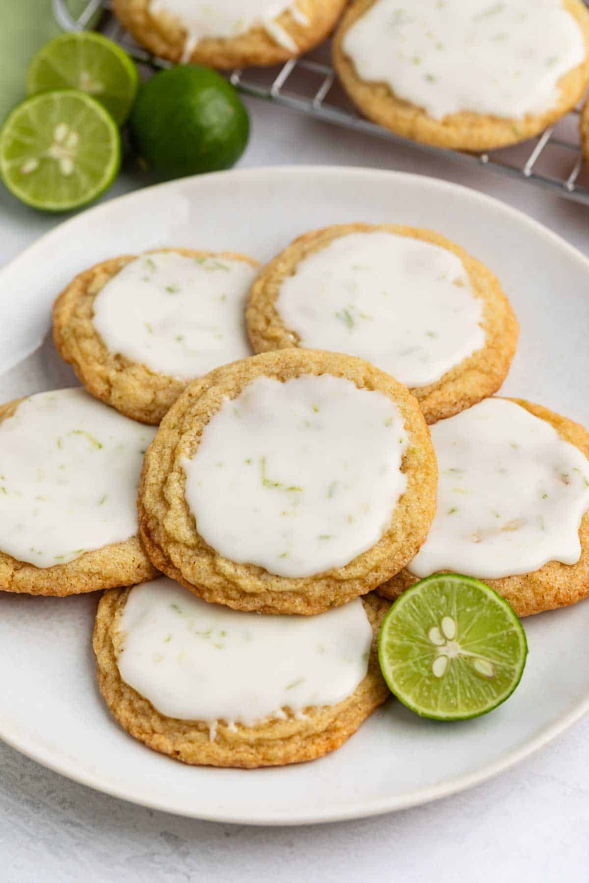 A plate of five cookies with white icing and lime zest, arranged around a sliced lime. More cookies and whole limes are in the background.