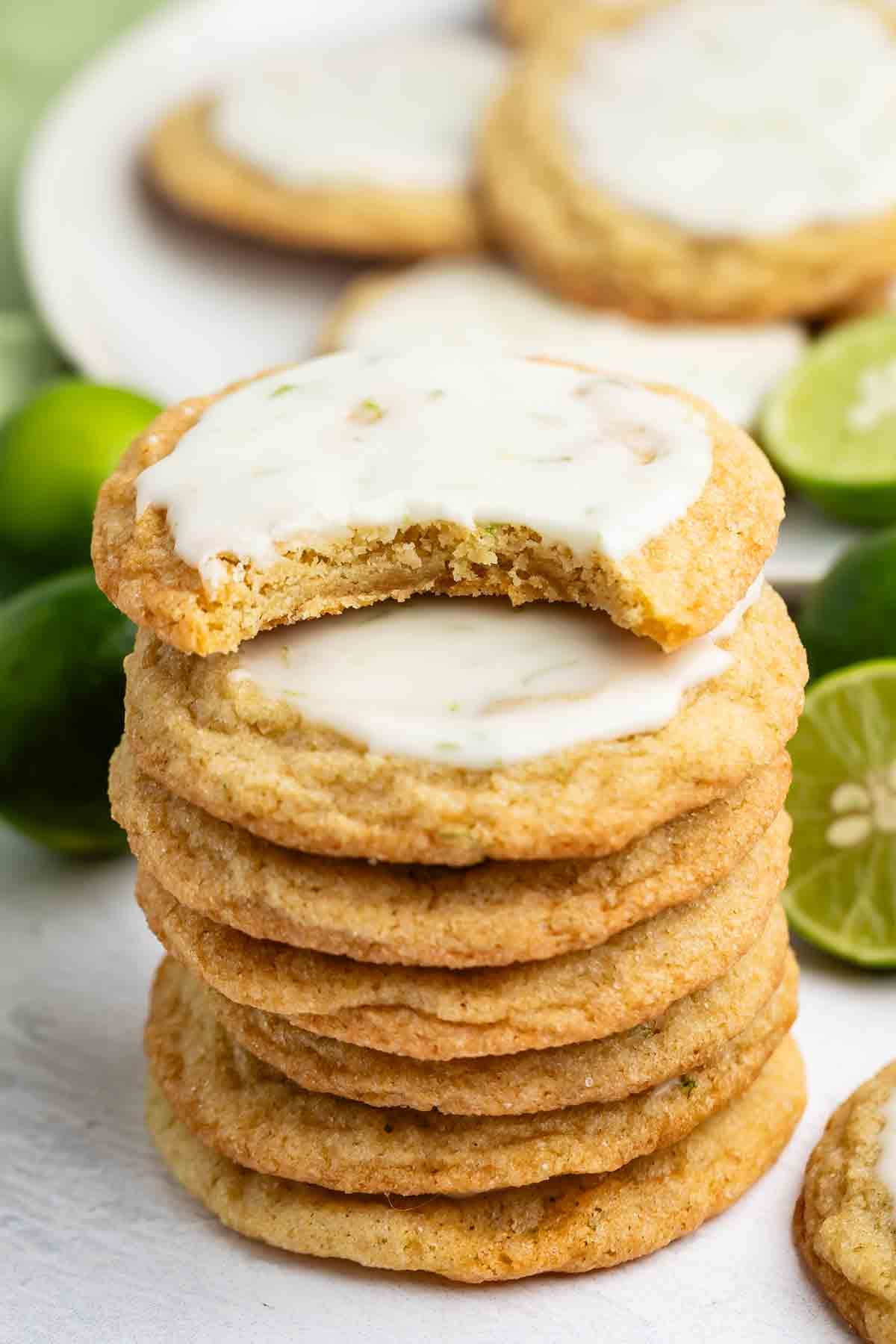 A stack of glazed cookies with one cookie on top that has a bite taken out of it. More cookies and halved limes are visible in the background.