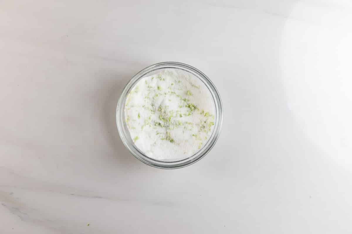 A small glass bowl filled with white sugar and scattered green zest, placed on a white marble surface.